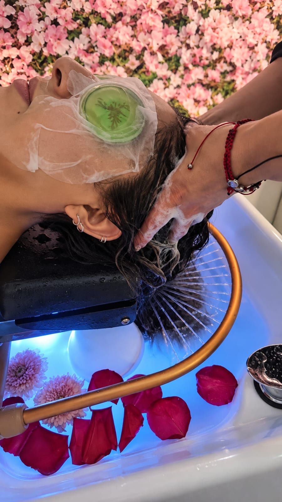 Clienta con antifaz verde recibiendo un masaje capilar entre flores rosadas y pétalos rojos